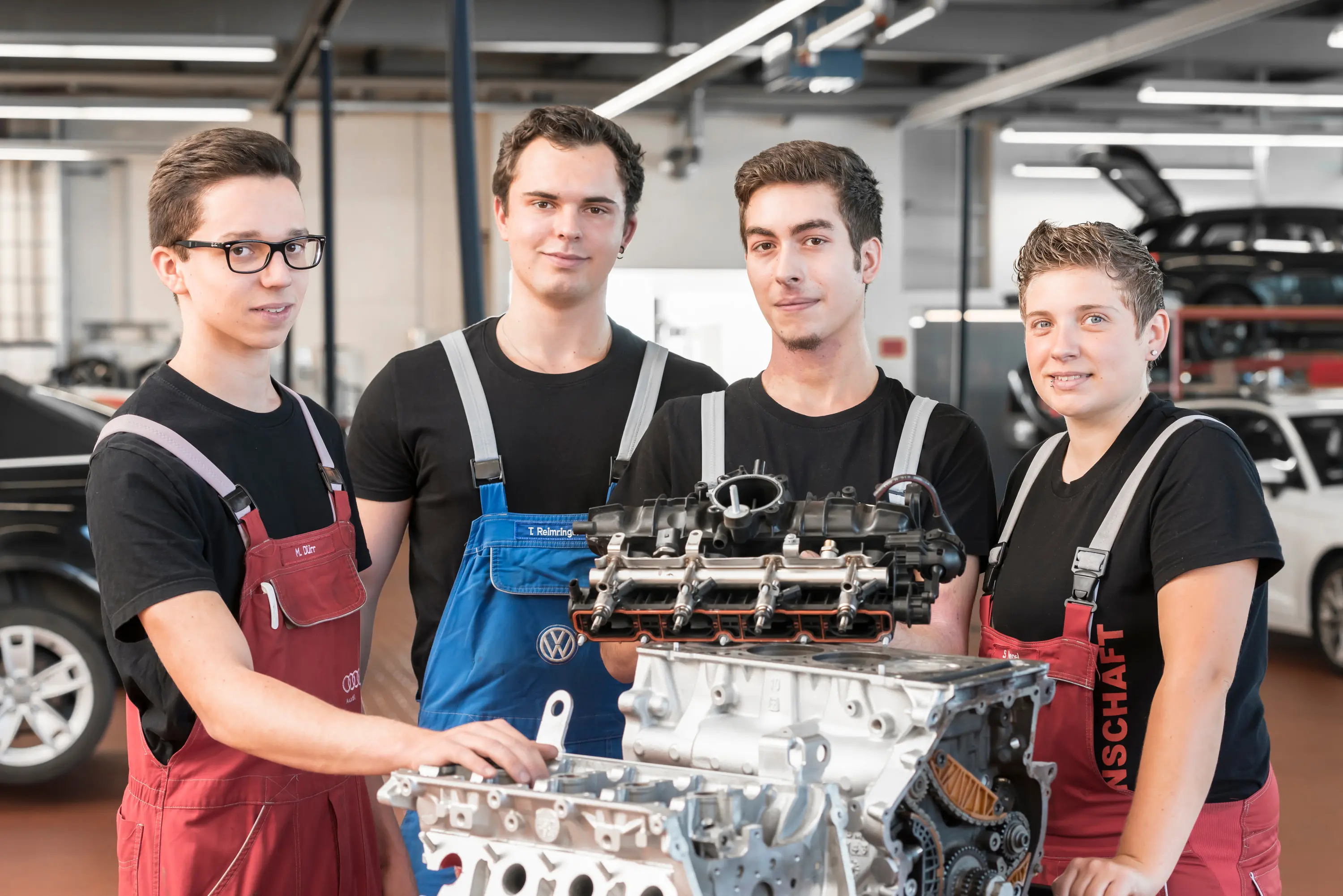 Vier Auszubildende in Arbeitskleidung an einem Motorblock im Autohaus